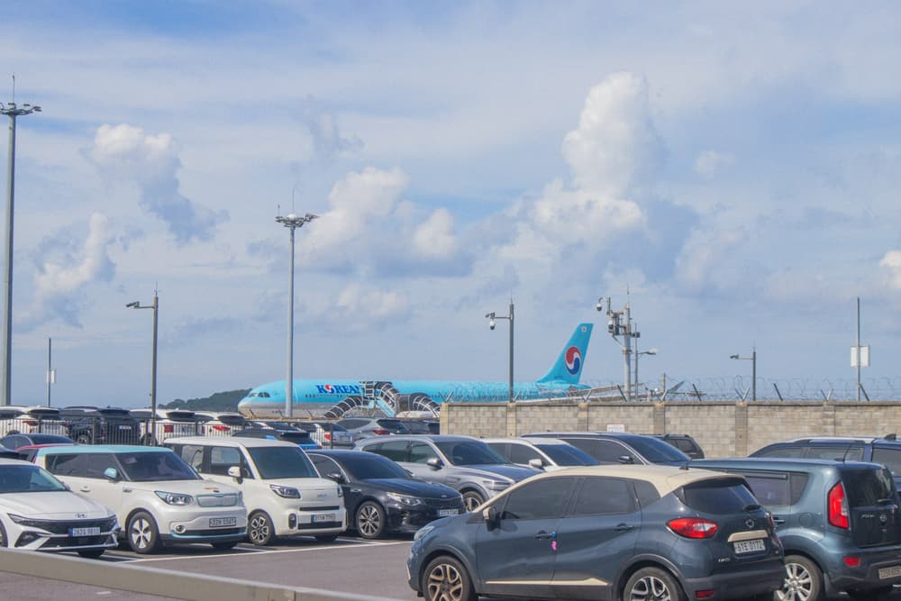 Parking przy lotnisku, w tle samolot startujący nad rzędami aut